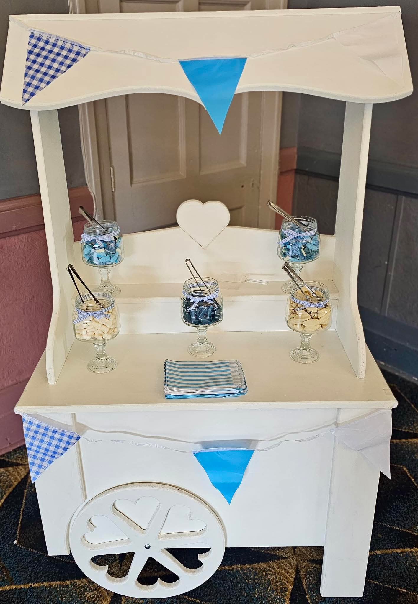 White sweet cart with heart cut-out wheel, dressed with blue gingham bunting and jars of sweets